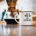 Morning Coffee - white and black life begins after coffee printed enamel cup beside 1/4 black liquid-filled glass flask on brown wooden panel