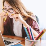 Student Loans - woman biting pencil while sitting on chair in front of computer during daytime